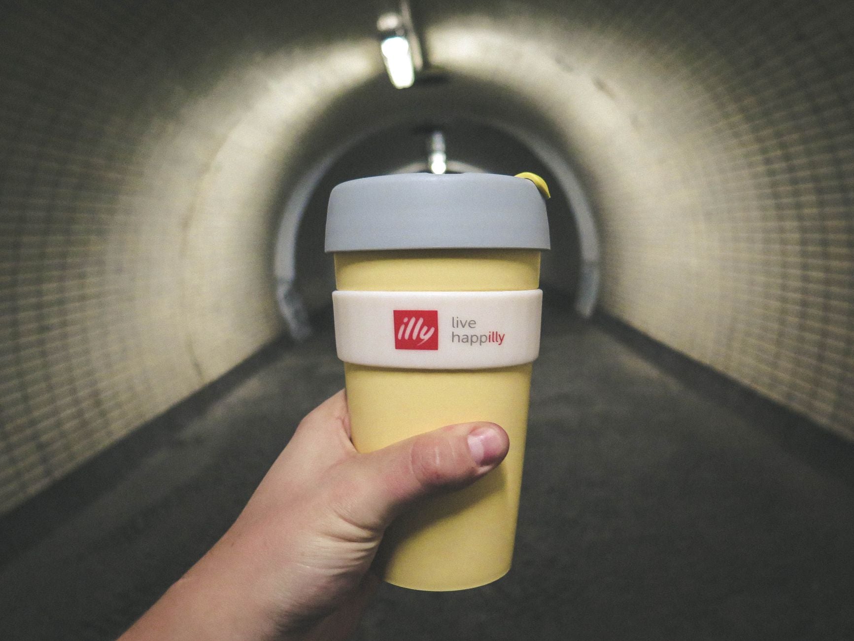Perspective shot of a hand holding a reusable coffee cup inside a dimly lit tunnel.