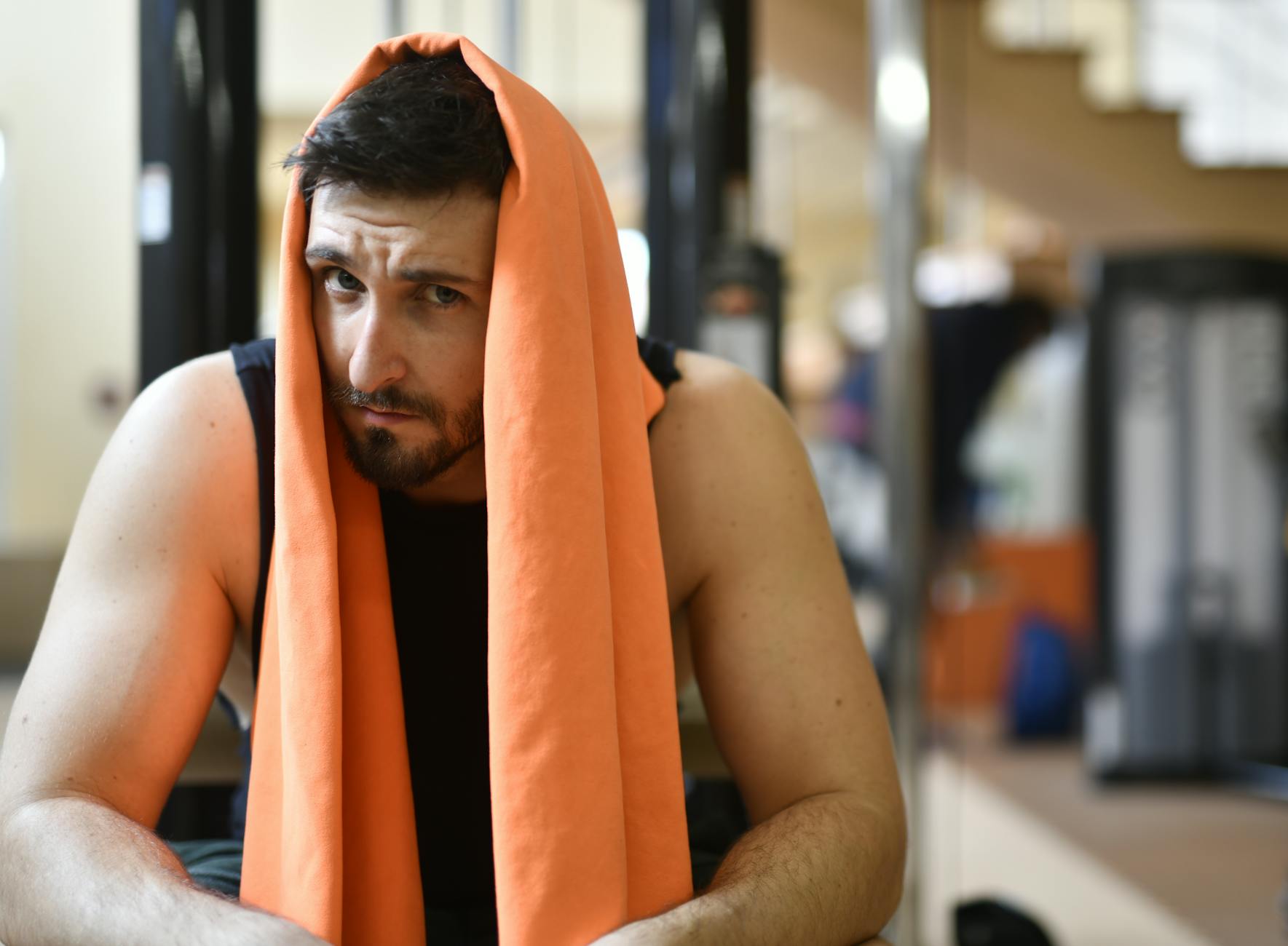 A tired man rests with an orange towel after a workout in the gym.