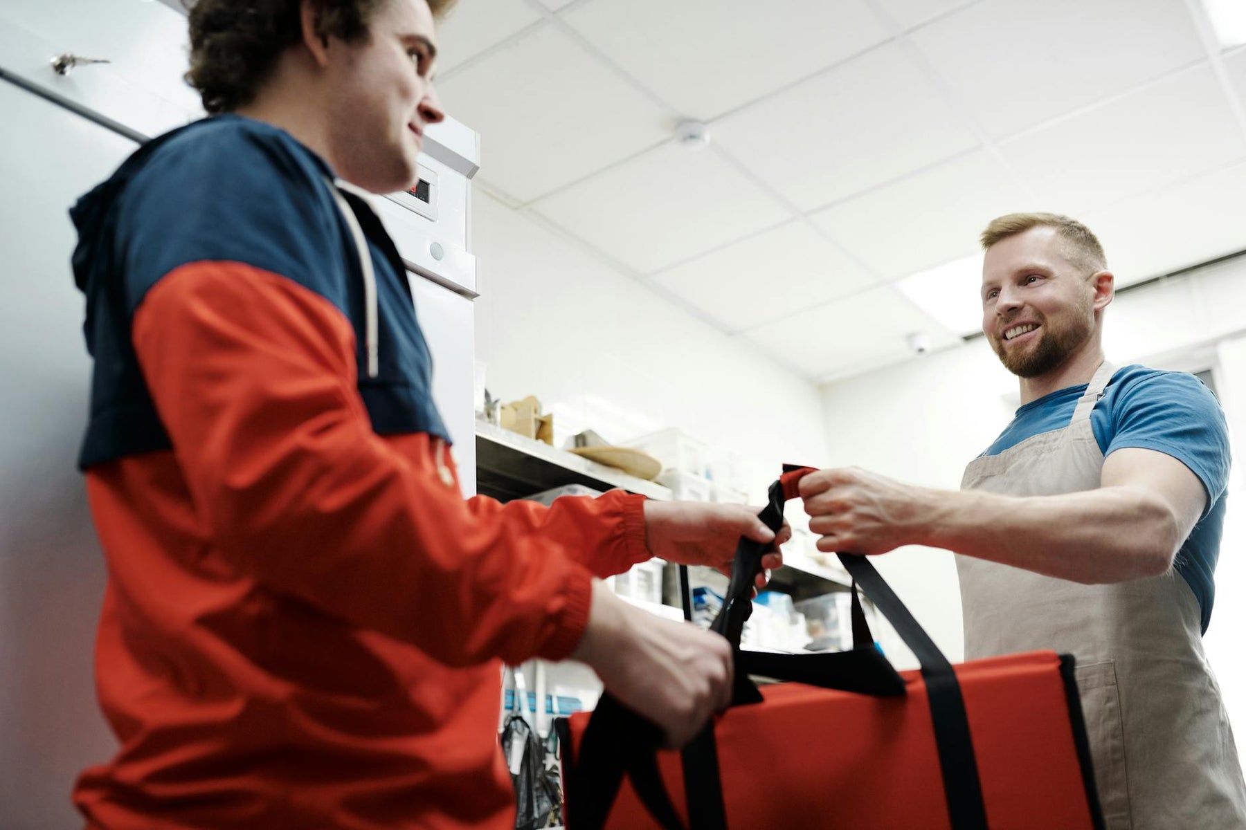 Smiling worker hands a thermal bag to a deliveryman in a busy kitchen.