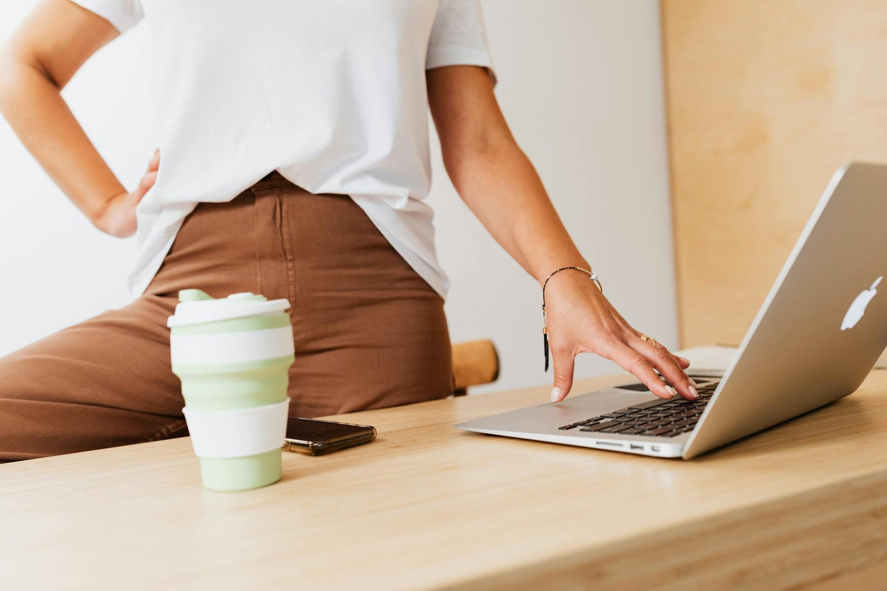 A woman stands by a wooden desk using a laptop with a reusable cup nearby. Modern lifestyle concept.