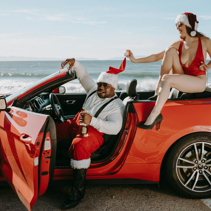 Santa Claus and woman in Santa hats enjoying a convertible by the seaside, showcasing a festive holiday scene.