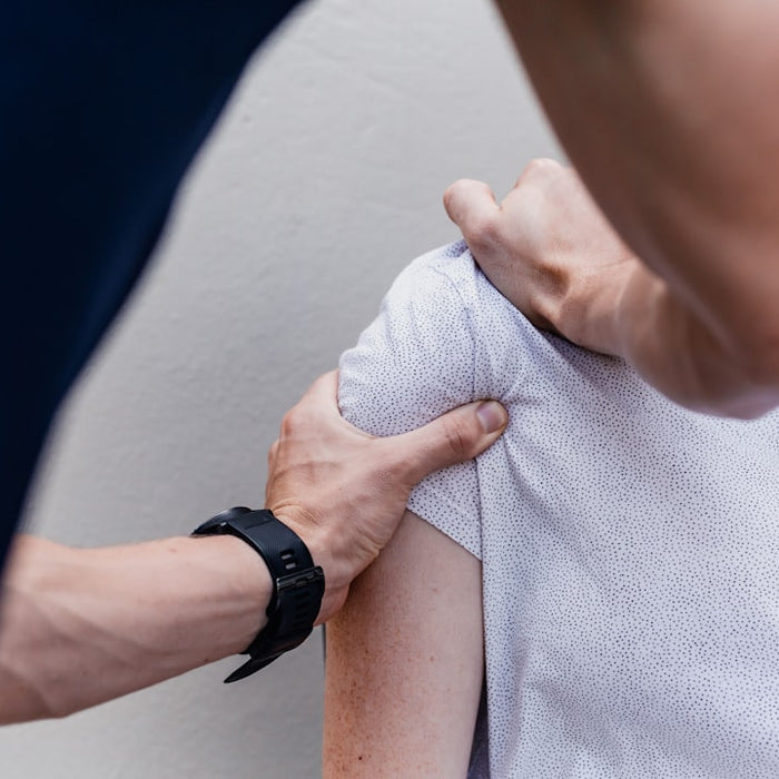 A person massaging the shoulder of another person wearing a white patterned shirt