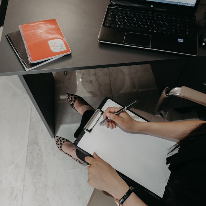 Person writing on clipboard at desk with laptop