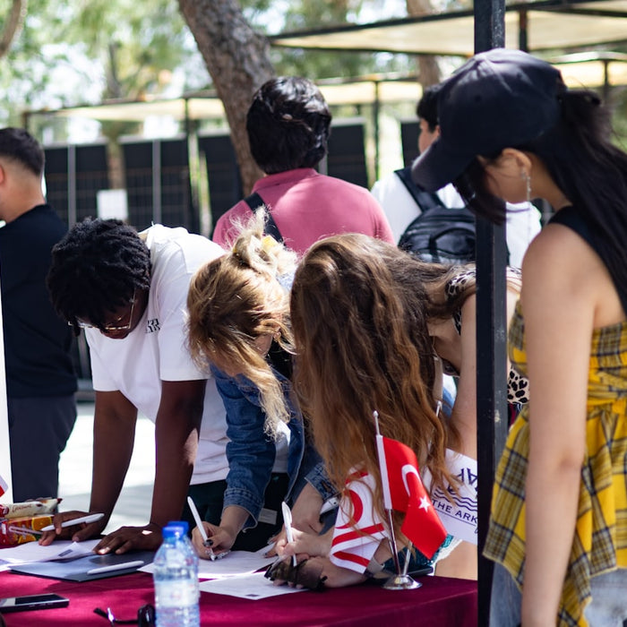 People signing papers at an outdoor table with flags.