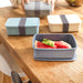 Seasons Dovi 800 ml Lunch Box - A set of three rectangular lunch boxes in blue, cream, and grey, with a grey strap, containing watermelon slices.