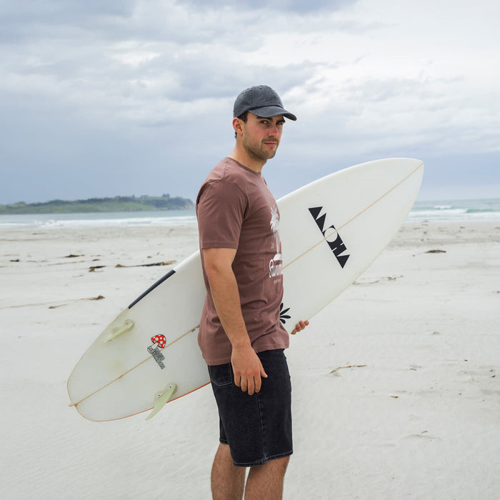 TRENDSWEAR Element Unisex T-Shirt - A man stands on a beach holding a surfboard, wearing a brown T-shirt and dark shorts.