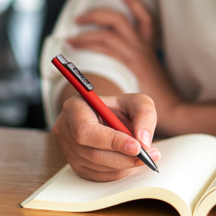 SPICE Slide Pen - A red metal pen held by a hand over an open notebook, featuring a branding logo on the pen.