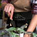 Keepsake Gardeners Set - A person using a black trowel with a wooden handle, wearing an apron with a logo, tending to a potted plant.