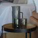 Keepsake Arlo Tumblers - Set of 4 - A drink bottle and two tumblers in smoky grey, placed on a round table beside a couch. The items feature a sleek design.