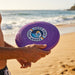 Aero Flyer - Large - A purple frisbee with a logo, held by a person at the beach, with waves and sand in the background.