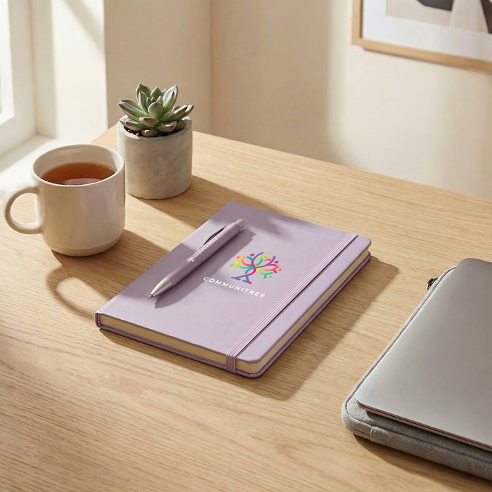 Omega Fashion Notebook and Pen Gift Set - A lavender notebook with a pen rests on a wooden desk, alongside a mug and a succulent plant.
