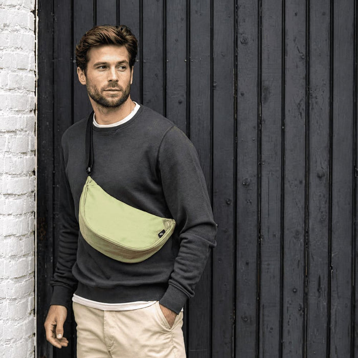 Byron Recycled Waist Bag 1.5L - A waist bag in light green worn across a man's chest, with a casual grey sweater and beige pants, against a dark wooden background.