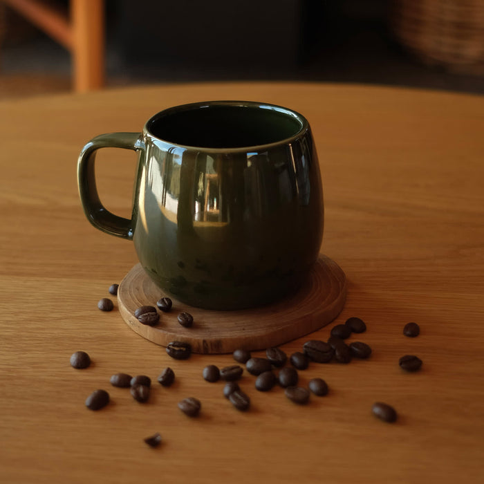 Keepsake Moda Coffee Mug – Set of 2 - A green ceramic coffee mug sits on a wooden coaster surrounded by coffee beans on a wooden table.