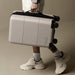 Kadi Carry-On Suitcase - Leisure - A grey carry-on suitcase with wheels being held by a person wearing white sneakers and ankle socks. The suitcase has a logo.