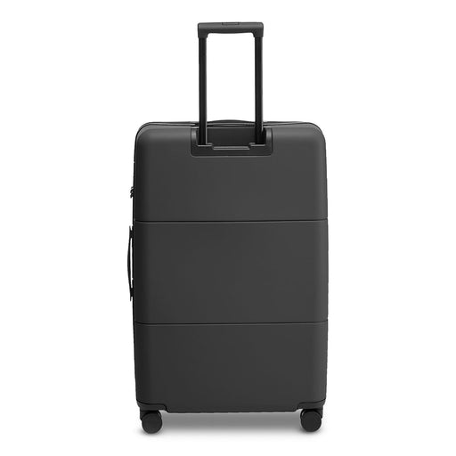 Kadi Check-In Suitcase - Large - A large black suitcase with a retractable handle and wheels, featuring a smooth surface and an attached logo.