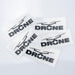 Prism Print - 40 x 20mm Sticker - Four black "DRONE" stickers on a clear backing, arranged on a light grey surface.