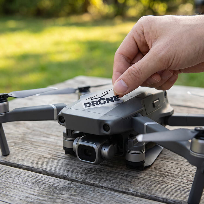 Prism Print - 40 x 20mm Sticker - A drone with grey and black components is being adjusted by a hand on a wooden surface outdoors.