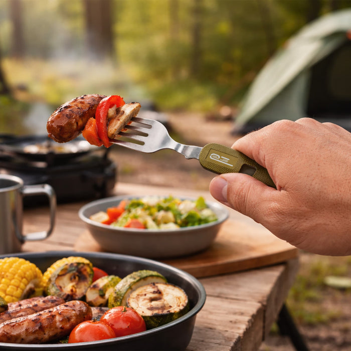 Camping Cutlery - A fork with a green handle holds grilled sausage and vegetables, with camping gear and food in the background.
