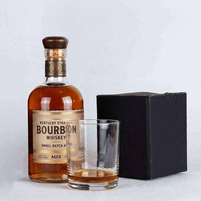 Rocks Glass - A bourbon whiskey bottle with a wooden cap, a rocks glass beside it, and a black box in the background.