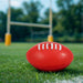 Mini Promotional Aussie Rules Ball - A red mini promotional Aussie Rules ball resting on green grass, with goalposts blurred in the background.