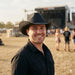 Airlie Paper Straw Hat - A man wearing a black straw hat smiles, with an outdoor festival stage and crowd in the background.