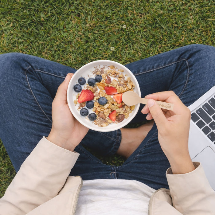 Pack of 100 Per Pack Disposable Birch Spoons/Forks (Small) - A person holding a bowl of granola topped with berries, using a small wooden disposable spoon, sitting on grass.