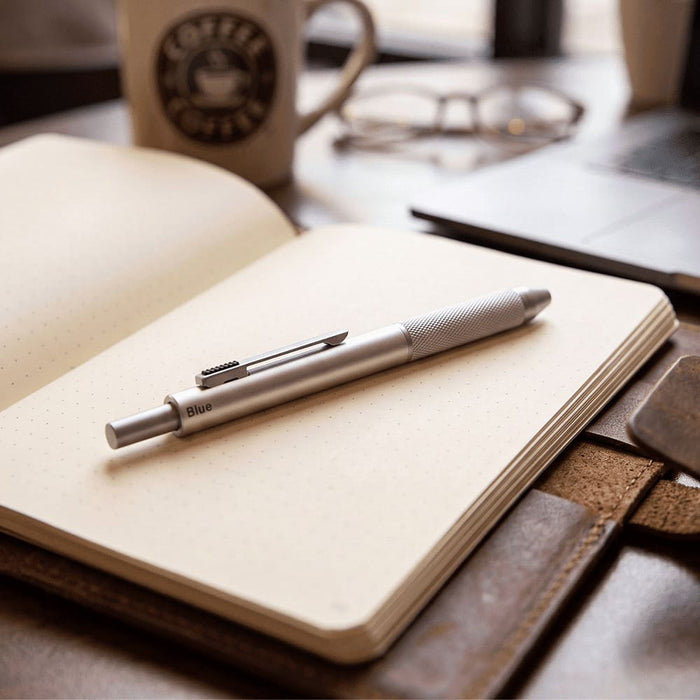 Tine 4-in-1 Gravity Recycled Stainless Steel Pen - A silver 4-in-1 pen rests on an open notebook, with a coffee cup and glasses in the background.