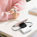 Avola Retractable Fast Charge Cable and Stand - A white retractable fast charge cable and stand connected to a smartphone on a desk, with a person writing in the background.
