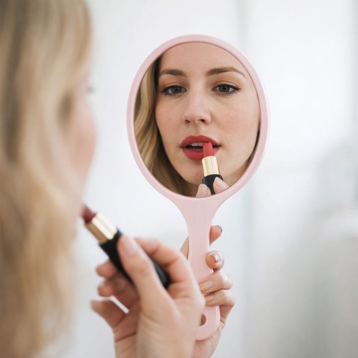 Chic Princess-Style Handheld Makeup Mirror - A pink handheld makeup mirror reflects a person applying lipstick, with a soft, neutral background.