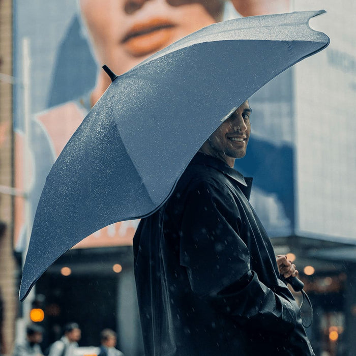 BLUNT Exec Umbrella - A navy BLUNT Exec Umbrella held by a person in a raincoat, with city buildings in the background.