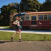 BLUNT Neo Umbrella - A woman carries a BLUNT Neo Umbrella in navy blue while walking beside a vintage tram in a green outdoor setting.