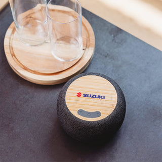 Boomer Bamboo Wireless Speaker - A round wireless speaker in black fabric with a bamboo surface on top, featuring a logo. Two clear glass cups are nearby.