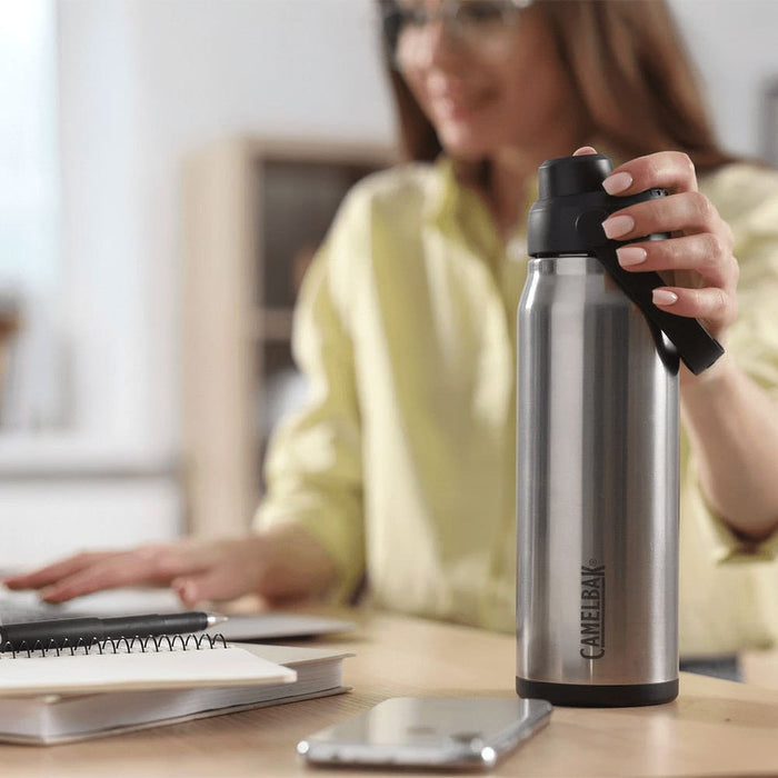 CamelBak - Thrive Chug VSS 0.75L - A drink bottle in stainless steel with a black cap, featuring a logo, placed on a desk next to a notebook and phone.