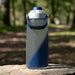 CamelBak - Thrive Chug 1.0L - A drink bottle in grey and navy blue, featuring a large handle and a spout, placed on a rock in a natural setting.