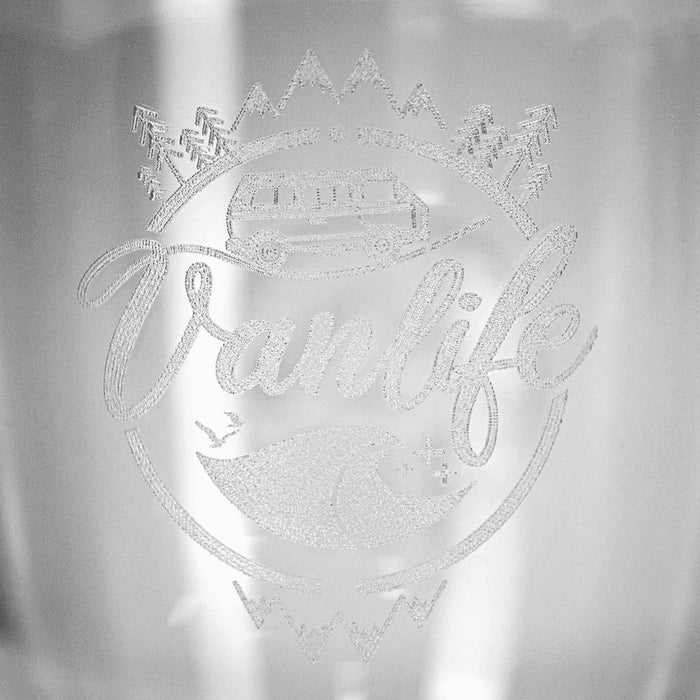 Glass Carry Cup - A glass cup with an etched design featuring a campervan, mountains, and waves, along with a logo.