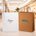 Paper Shopping Bag - Two paper shopping bags are displayed, one white with a logo and the other brown with a logo, set in a retail environment.