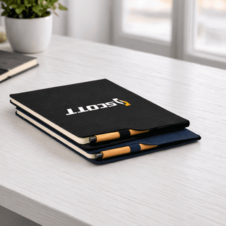 SunScribe Notebook (A4) - A black and navy A4 notebook with a logo, stacked on a desk next to a small pot plant.
