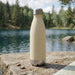Komo Wheat Straw Drink Bottle - A drink bottle in a light beige colour with a stainless steel base, set against a scenic lakeside background.