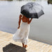 Poppins Umbrella - A black umbrella with a logo held by a person in a white dress, standing by the water.