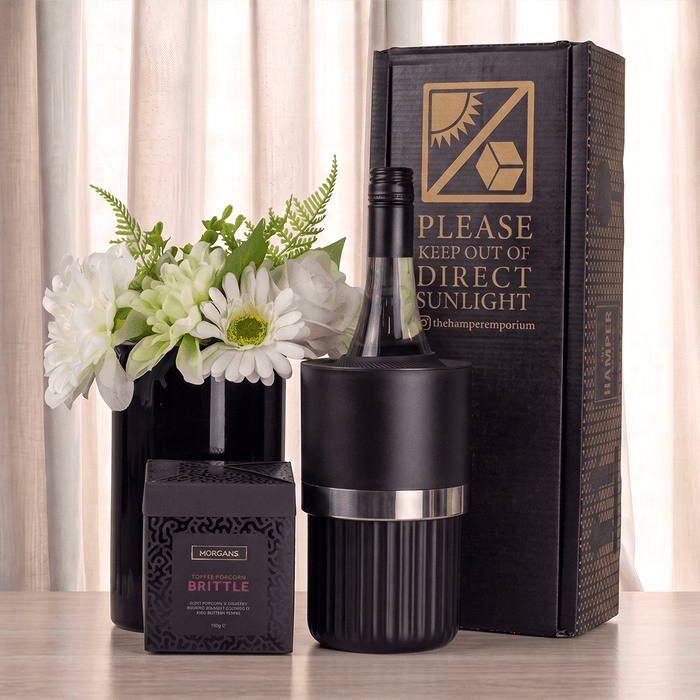 The Entertainer Set - A black wine bottle cooler with silver band, a box of brittle, and a vase with white flowers, arranged on a table.