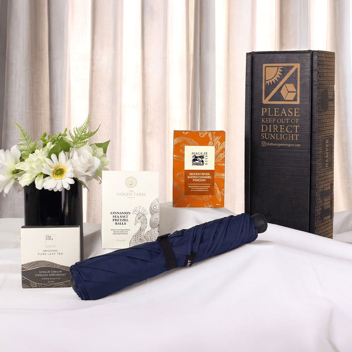 The Blunt Umbrella All Weather Set - A navy umbrella, surrounded by boxes and packets, set against a light backdrop with a vase of flowers.