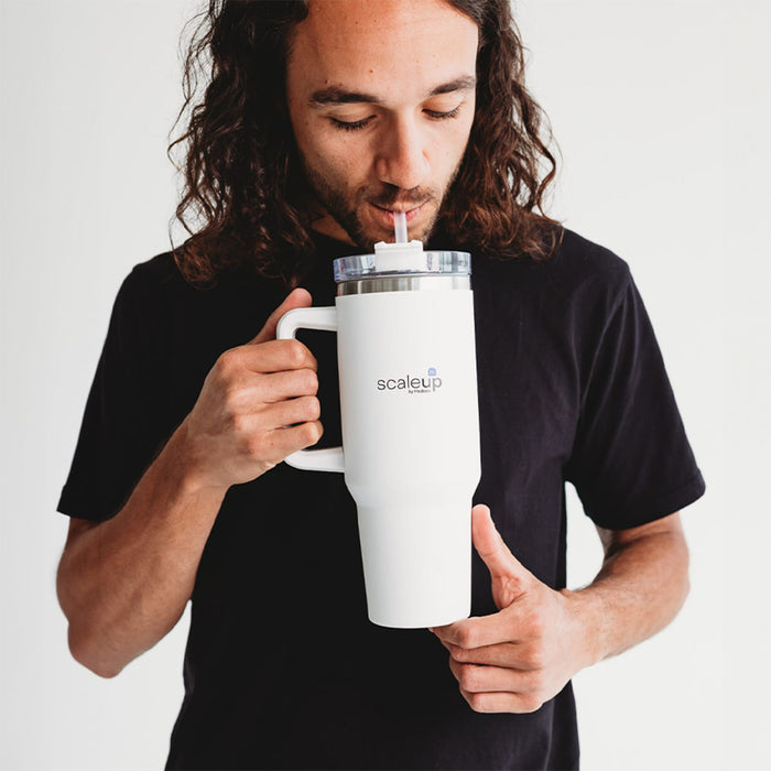The Hippo 1.2L Mug - A drink bottle in white with a transparent lid and straw, featuring a logo, held by a person.