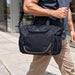 High Sierra Crib Bag - A navy cooler bag with a sturdy strap is being carried by a person walking on a city sidewalk. It has a logo.