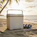 13L Cooler Box - A white 13L cooler box on a sandy beach, accompanied by a blue-striped towel and sunglasses.