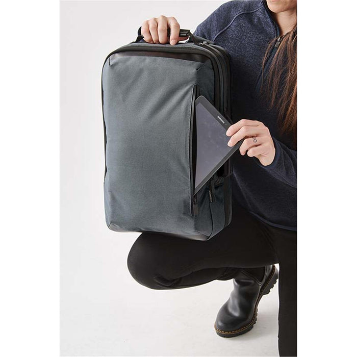 Hedmark Commuter Backpack - A person holds a grey backpack while placing a tablet into a front pocket. The backpack has a logo.