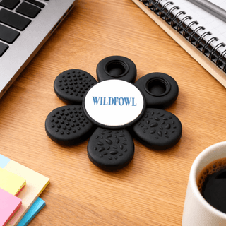 The Spinzy - A black fidget spinner in a flower shape with a central logo, placed on a wooden desk near a coffee cup and stationery.
