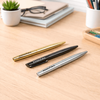 The Rhi Metal Ball Pen - Three metal ballpoint pens in gold, black, and silver arranged on a wooden desk with stationery in the background.