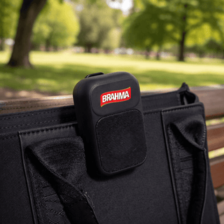 The Rebel Clip Speaker - A black portable speaker with a logo, attached to a bag, set against a green park background.