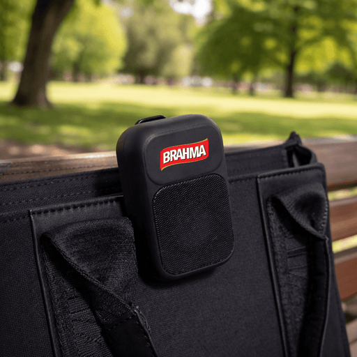 The Rebel Clip Speaker - A black portable speaker with a logo, attached to a bag, set against a green park background.