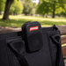The Rebel Clip Speaker - A black portable speaker with a logo, attached to a bag, set against a green park background.